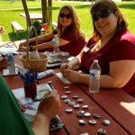 Katie Radford and Katie (Koch) Code sit at a picnic table with Augsburg buttons and other swag